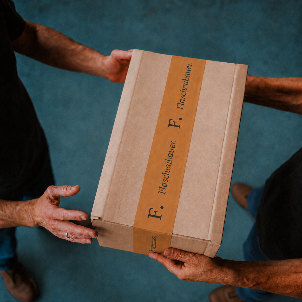 Zwei Personen übergeben ein braunes Versandpaket mit bernsteinfarbenem Klebeband und großem Aufdruck „F. Flaschenbauer.“ vor einem blaugrauen Hintergrund.