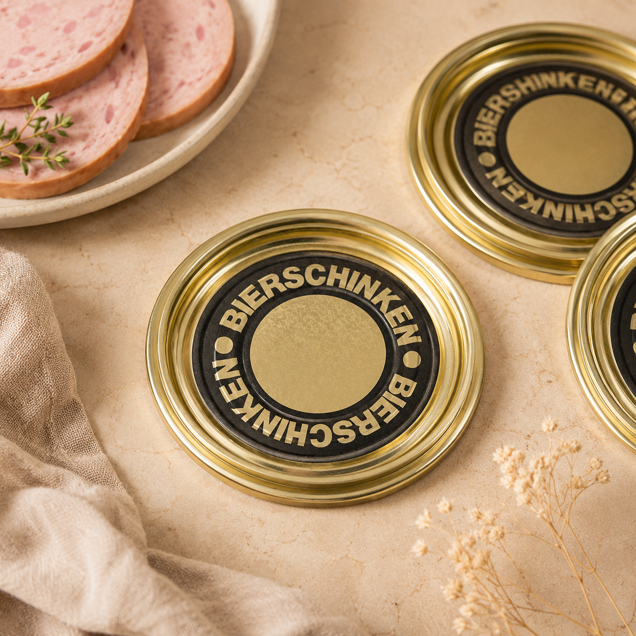 Goldene Falzdeckel mit der Aufschrift „Bierschinken“ als Flatlay auf beigem Untergrund, dekoriert mit Bierschinkenscheiben und Leinentuch.